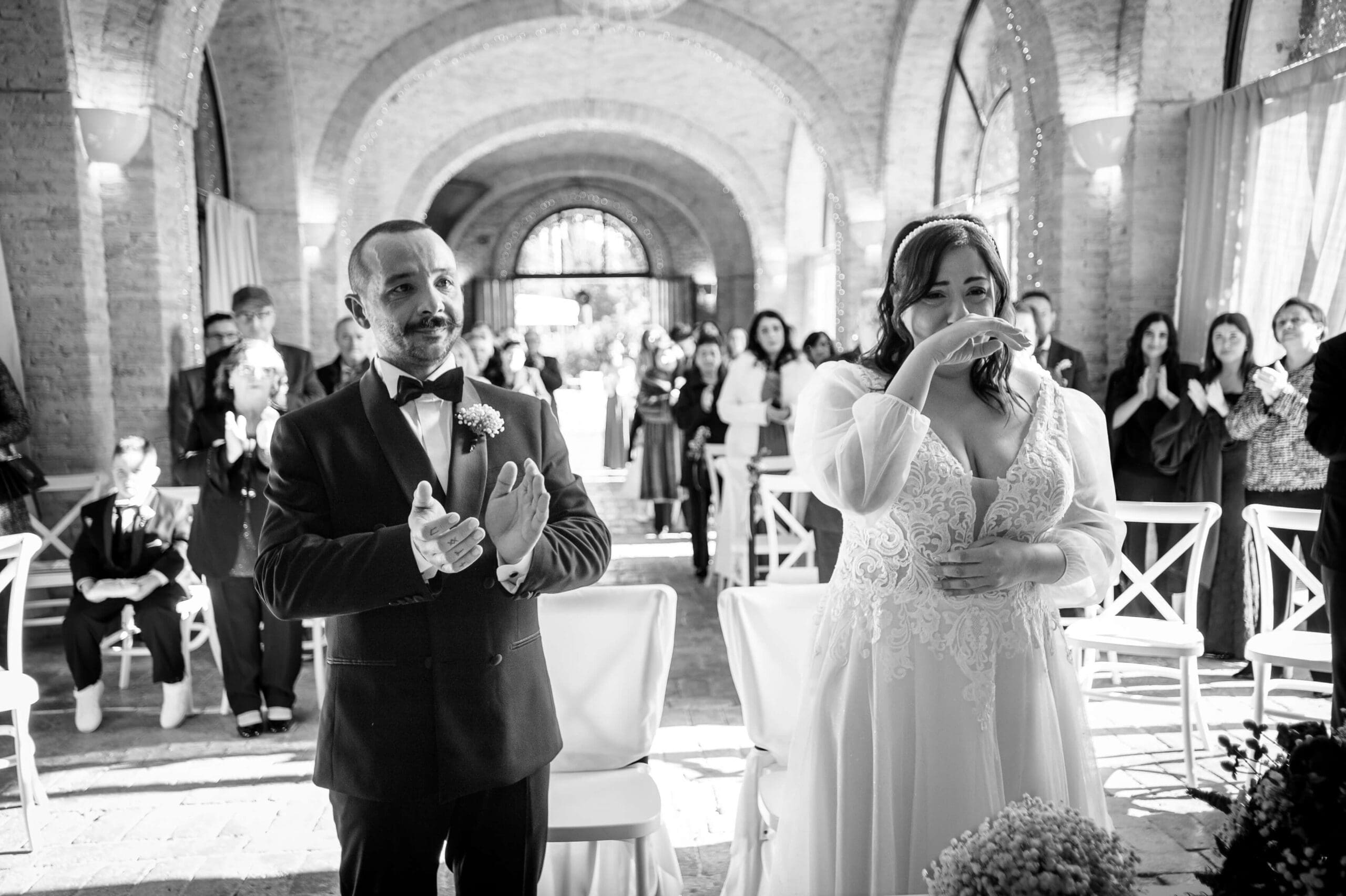 Un momento di forte commozione durante la cerimonia: la sposa si asciuga una lacrima mentre lo sposo applaude accanto a lei, circondati dal calore degli invitati in una sala suggestiva.